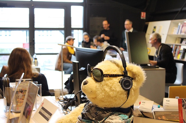 Café musique de décembre, en premier plan un ours peluche avec des lunettes solaires et du monde en arrière plan.