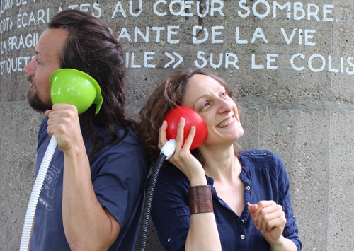 Deux adultes, un homme et une femme, tenant chacun un tuyau muni d'un entonnoir et le porte à son oreille