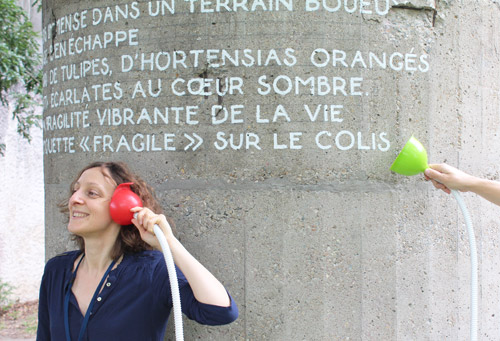 Une jeune femme tient en souriant un entonnoir avec un tuyau au niveau de son oreille et derrière elle un mur contenant un texte peint en blanc