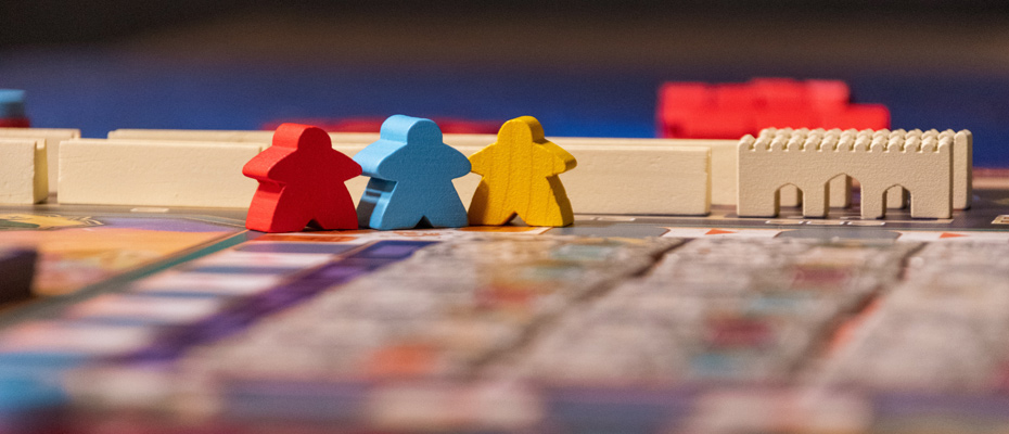 Gros plan sur un plateau de jeu de société avec trois pions en bois (personnages rouge, bleu et jaune) et un château en bois