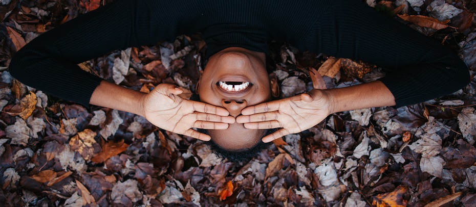 femme allongée sur des feuilles mortes, souriante et en mettant ses mains devant ses yeux