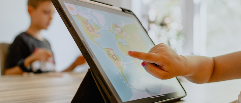 Une main d'enfant devant un écran de tablette et un deuxième enfant en arrière plan (flou)