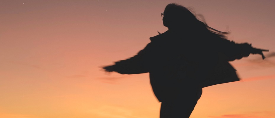 Vue à contre jour (soleil couchant) sur une jeune femme qui lève les bras comme pour voler