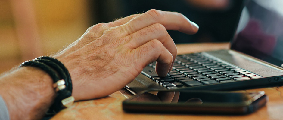 Une main masculine tapant sur le clavier d'un ordinateur portable posé sur une table. Un smartphone posé à côté.