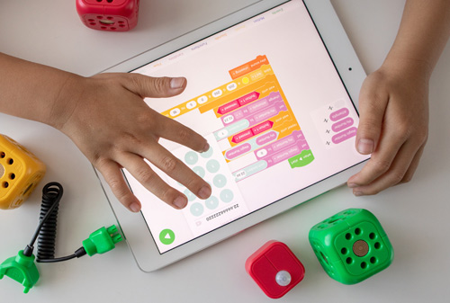 une main d'enfant sur une tablette blanche avec à l'écran des cases de couleur jaune, rouge, verte.