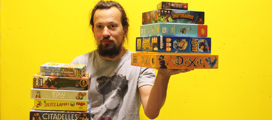 Jeune homme souriant tenant une pile de boîtes de jeux de société dans chaque main, sur fond jaune vif