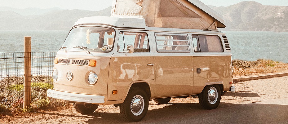 Un van vintage couleur beige et blanc avec une tente sur le toit du véhicule. Stationné sur un chemin avec la mer au fond