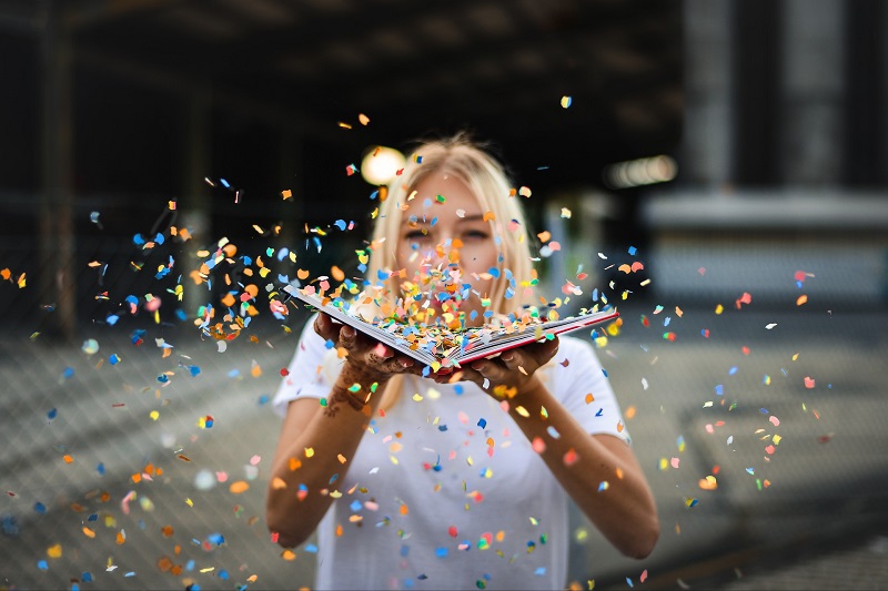 femme soufflant sur un livre d'où s'envolent des confettis multicolores