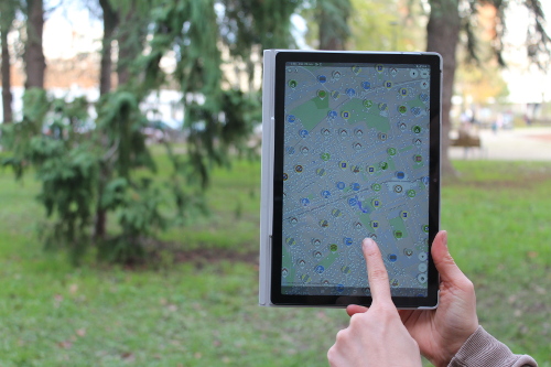 Main pointant du doigt une tablette affichant l'application Streetcomplete. Arrière plan : pelouse et arbre.