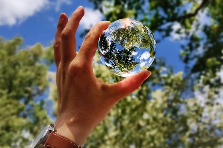 Une main vue en contre plongée tenant une boule de verre dans laquelle se reflète la cime d'arbres en arrière plan avec un ciel bleu rappelant la planète terre.